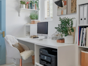Compact white built-in home office desk with shelves and printer storage, styled with plants and files; bright small-room workspace near a window.